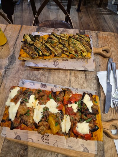 The image shows two rectangular pizzas on wooden serving boards, placed on a wooden table. The top pizza is topped with grilled zucchini slices and herbs. The bottom pizza is topped with grilled eggplant, bell peppers, tomatoes, and melted white cheese. A fork and knife are placed to the right of the bottom pizza. The background shows a glimpse of chairs and a wooden floor, suggesting an indoor dining setting, possibly a restaurant. The pizzas are presented on patterned paper liners. There are no people visible in the image. Based on the text on the paper liners, which includes "Aroma Sourdough" and "Cambridge United Kingdom", this scene is likely located in Cambridge, United Kingdom.