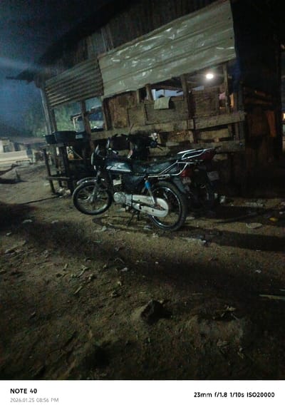 A dimly lit scene features two motorbikes parked in front of a wooden and corrugated metal structure. The ground is uneven and covered in dirt and debris. A dim light illuminates the building's interior, highlighting its makeshift construction. The overall atmosphere is dark and gritty, suggesting a rural or impoverished setting. The date, time, and camera settings are displayed at the bottom of the image.