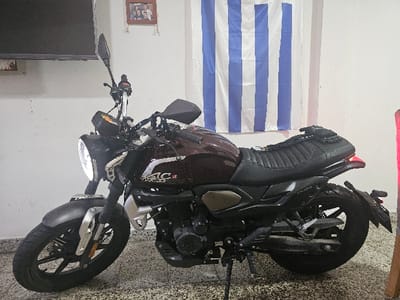 A dark-colored motorcycle sits indoors on a tiled floor, its headlight illuminated. The bike features a brown gas tank with silver lettering, a black seat with a textured surface, and black wheels. Behind it is a striped flag of white and blue, vertically displayed. A television is visible in the upper left corner, and a framed photo hangs on the wall above it.