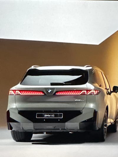 A rear-view, medium shot captures the back of a silver BMW X5 parked indoors. The car is positioned centrally, facing away from the viewer. The vehicle's taillights are illuminated, emitting a red glow. A license plate frame displaying "BMW X5" is affixed below the taillights. The interior environment appears to be a studio or showroom, with plain, light-colored walls and flooring. The lighting is soft and diffused, casting subtle shadows. There are no people or other vehicles visible in the frame. The location of Sant'Angelo di Piove di Sacco, Italy, is not ascertainable from the image.