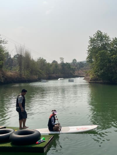 The image shows a scene on a body of water, likely a lake or river, in Tambon Nam Phrae, Thailand. The water is greenish-brown and calm, with light reflecting off the surface. There are trees and vegetation along the banks of the water. In the distance, there are wakeboarding obstacles set up on the water.
Two people are visible on a floating platform. One person is sitting on a paddleboard, wearing a life vest and a visor. They appear to be preparing to paddle. The other person is standing on the platform, dressed in shorts and a shirt, looking at the person on the paddleboard. Two black inner tubes and a water gun are on the platform. Powerlines can be seen in the background.