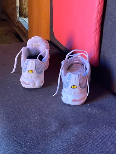 In Borodulino, Russia, a pair of white Nike Air Max Plus TN sneakers are placed on a dark blue, textured surface, likely upholstery. The shoes are positioned with their backs facing the camera, and their laces are loosely tied. A bright pink, rectangular object is visible in the background, partially obscuring a wooden structure. The lighting suggests an indoor environment.