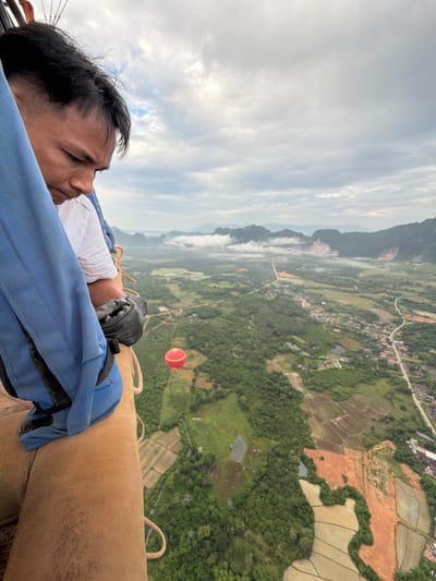 A man, wearing a white shirt and black gloves, is in a hot air balloon basket looking down at the landscape. The basket is made of brown material and has blue straps. Below the balloon, a green and red hot air balloon is visible, along with a village and surrounding farmland, including rice paddies. Mountains are visible in the distance, partially covered by clouds. The location is identified as Hong Ngern Village, Laos.