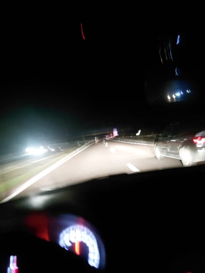 A blurry, dark night scene possibly taken from inside a moving vehicle. A speedometer glows on the right. Streaks of light suggest motion, with blurred headlights of oncoming traffic and streetlights visible. A dark expanse on the left hints at the landscape outside. The overall impression is of speed and nighttime travel.