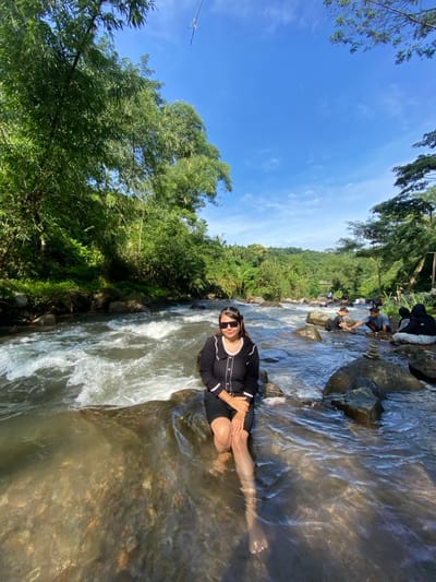 A woman in a black dress and sunglasses sits on a rock in a river, with her legs submerged in the water. Other people are seen in the background, sitting on rocks near the riverbank. Lush green trees and vegetation surround the river, and a clear blue sky is visible above. The water is flowing rapidly, creating white water near the woman.