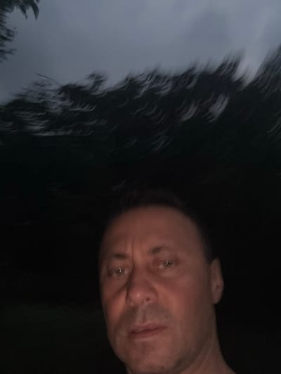 The image shows a close-up, blurry selfie of a man's face taken at night. The man appears to be outdoors, as indicated by the dark, out-of-focus foliage in the upper portion of the image. His face is illuminated by a light source, possibly from the camera, casting shadows and highlighting his features. The man's expression is neutral. The overall quality of the image is poor due to low light and motion blur. There is no indication of the specific location within the city of Tandzia, Georgia, or any contextual details to suggest the activity or purpose of the photograph.