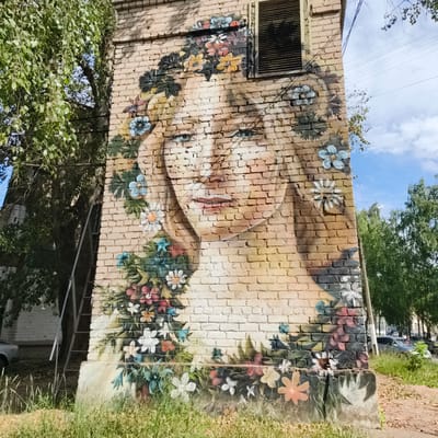 The image displays a mural of a woman's face adorned with flowers, painted on the side of a brick building in Chaikovsky, Russia. The mural covers a significant portion of the building's facade, which appears to be part of a residential structure given the presence of a ventilation grate near the top. The woman's face is rendered in soft, pastel tones, with blue eyes and a gentle expression. The flowers depicted are a mix of colors and types, with some appearing as stylized daisies and others as more abstract floral shapes. Green foliage is integrated into the floral elements, creating a natural and organic feel.
The building itself is constructed of light-colored bricks, and the mural is painted directly onto this surface. To the left of the mural, a large tree with green leaves casts shadows on the wall, partially obscuring the artwork. A metal ladder leans against the building next to the tree, suggesting it may have been used for the mural's creation or for maintenance. The ground in front of the building is covered with grass and some dirt or gravel.
In the background, more trees and a clear blue sky with scattered clouds are visible. Power lines are strung across the sky. To the right of the building, parked cars and a glimpse of a street suggest an urban or suburban setting. The overall impression is one of an urban landscape enhanced by vibrant street art.