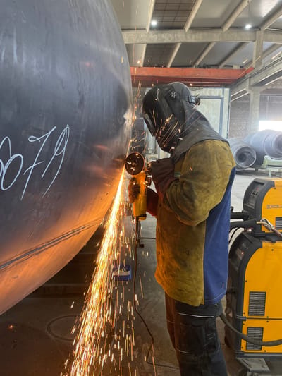 An industrial worker is depicted using an angle grinder on a large, curved metal object, likely within a fabrication workshop in Tomba, Montenegro. The worker is positioned towards the left of the frame, facing right, and is wearing a dark welding helmet, heavy-duty protective gloves, and a two-toned protective jacket or apron in blue and yellow-brown.
The primary activity is metal grinding, evident from the copious, bright orange sparks emanating from the point of contact between the yellow DeWalt angle grinder and the dark metallic surface. These sparks create a striking visual, extending across the mid-ground and foreground of the image. The large metal object being worked on appears to be a section of a pipe or a spherical tank, featuring a seam line and white chalk markings, which are partially legible as "170" or "174."
In the background, an industrial setting is visible with concrete structural elements, metal framework, and what appears to be large machinery. A prominent yellow and black industrial machine is positioned above the worker, displaying text such as "KOLAY" or "KOLAR" and "IGBT," suggesting it is a piece of welding or power supply equipment. The overall scene conveys an active, manufacturing environment. The lighting suggests an indoor setting, likely during daytime hours, with no discernible weather effects. The worker's posture and focus indicate a concentrated and precise task.