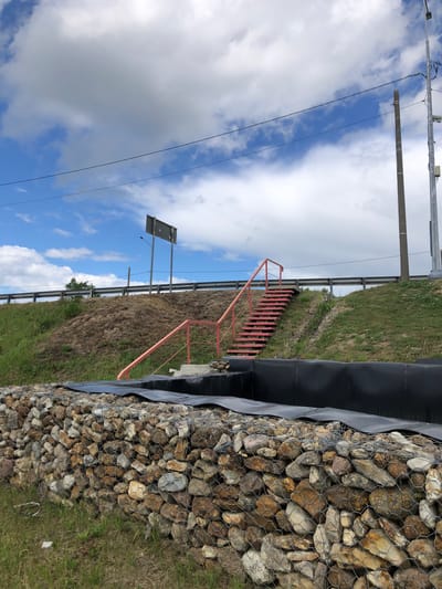 The image depicts an outdoor scene featuring a gabion wall constructed from various stones and wire mesh in the foreground. Behind the wall, there is a black plastic liner, possibly for a pond or water feature, and a set of red metal stairs leading up a grassy slope. The slope is covered with dirt and grass, and a guardrail is visible at the top, suggesting a road or highway nearby. A tall metal pole with what appears to be a camera and other equipment is situated on the right side of the frame, and a signpost with a blank sign is visible on the left. The sky is blue with scattered white clouds. The overall setting appears to be an undeveloped or construction area. Based on the user's prompt, the location is identified as Nadezhda, Russia. There are no people visible in the image, and no actions or interactions are taking place.