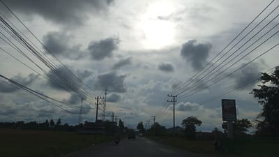 A cloudy, overcast sky with the sun partially visible through a break in the clouds is the dominant feature of this daytime, outdoor scene. A road stretches into the distance, lined with telephone poles and power lines. On the left side of the road, there are houses and trees, with a field in the foreground. On the right side, there are more trees, houses, and a billboard advertising "COKLAT" (chocolate). A few vehicles are present on the road, including a motorcycle with a rider in the foreground on the left, and several cars further down the road. The overall atmosphere is somewhat somber due to the heavy cloud cover. This scene is located in the city of Cotbadatunong, Indonesia.