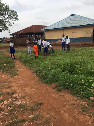 The image shows a group of people, mostly children, standing near buildings in an outdoor environment in Enyibichiri, Nigeria. The buildings have corrugated metal roofs, and one has faded paint on its walls. The children are wearing various outfits, including white shirts and blue skirts or trousers, and some wear orange or other colored shirts. A dirt path is visible in the foreground, with grass and weeds growing nearby. The sky is overcast. The image provides no further information about specific city locations.