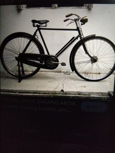 A vintage black bicycle with a sprung saddle is propped up on a stand on a cracked concrete surface. The bicycle has a headlight mounted on the handlebars and a chain guard. The background is a textured wall, possibly of concrete or brick. The scene appears to be outdoors. A dark rectangular frame at the bottom of the image contains text, some of which is legible. The legible text reads "LAPAK BARANG ANTIK" and "antik, barang koleksi, jam...". The context of "Kualaraja, Indonesia" is not directly discernible from the image itself, but the text on the frame suggests a marketplace or shop selling antique items, which could be located in such a place.