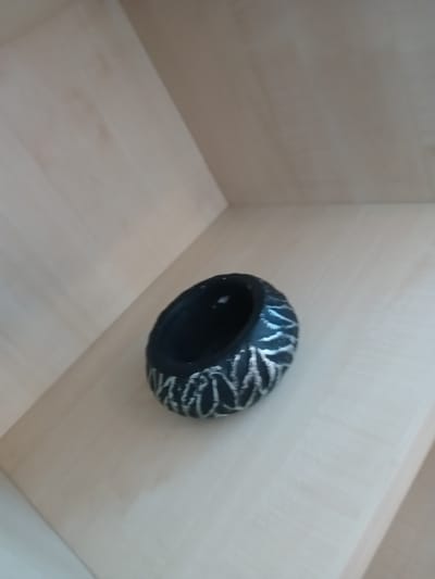 A dark, circular object with a textured pattern sits on a light-colored wooden shelf. The object appears to be a decorative bowl or holder, with a matte black interior and a textured exterior featuring a repeating silver pattern that resembles stylized leaves or waves. The shelf is made of light wood with visible grain and is positioned at an angle, suggesting it is part of a tiered shelving unit. The background is out of focus and consists of more of the same light-colored wooden shelving. No people are visible in the image, and there are no indications of the object's function beyond its decorative appearance. The scene is indoors, and the lighting appears to be ambient, casting soft shadows. The location is not specified in the image, and therefore, its presence in Ankara, Turkey, cannot be confirmed.