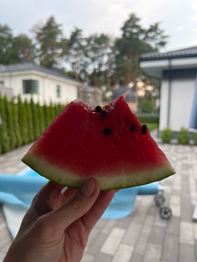 A hand holds a slice of watermelon with a bite taken out of it. The watermelon is red with black seeds and a green rind. In the background, there are houses, trees, and a cloudy sky, indicating an outdoor setting. The environment appears to be residential, possibly in a suburban area. The overall scene suggests a moment of casual enjoyment of a summer treat in an outdoor space. The specific location is in Briukhovychi, Ukraine.