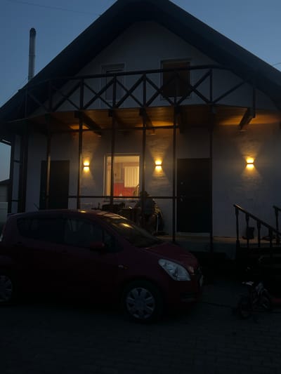 This media file depicts a nighttime scene in Solontsy, Russia, featuring a red car parked in front of a modern house with an A-frame design. The house is illuminated by warm outdoor lights, casting a glow on its white facade and wooden beams. A chimney is visible on the left side of the image, and a staircase with dark railings leads up to the house entrance. The sky is dark blue, suggesting a clear night. The overall mood is calm and serene, with the lighting creating an inviting atmosphere. No people or specific activities are visible, but the presence of a parked car and the lit house suggest a residential setting. The ground appears to be paved with cobblestones.