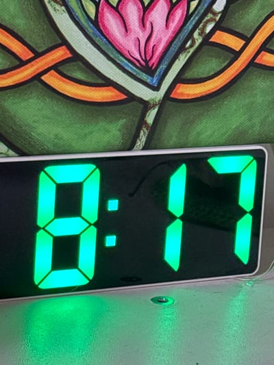 The image depicts a digital clock displaying the time 13:08. The clock has a black background with green LED digits. To the left of the clock is a portion of a patterned fabric with a predominantly green background, featuring orange and pink design elements. To the right of the clock is a plain, light-colored surface with a slight texture. The environment appears indoors with ambient lighting. There is no indication of location in Ketoshi, Russia or any other city.