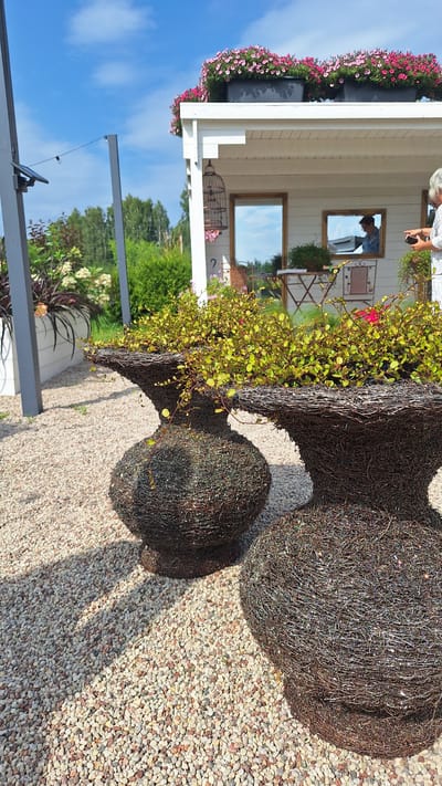 The image depicts a charming outdoor setting with two decorative planters in the foreground, constructed from dark, interwoven wire and filled with lush green trailing plants. These planters are placed on a bed of small, light-colored gravel. In the background, a white, open-sided structure resembling a shed or small pavilion is visible. This structure is topped with rows of pink and magenta flowers in planters, and has a decorative birdcage hanging from its overhang. Two framed mirrors hang on the wall of the structure, reflecting the blue sky and trees. A woman in a white shirt is visible in the right background, holding a dark object and looking towards the structure. The environment suggests a garden or a recreational area, likely a private garden or a small business in a rural or suburban setting. The specific location within Flokši, Latvia, is not identifiable from the visual information provided in the image.