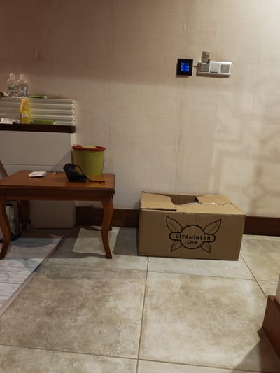 This is an indoor scene, likely a room in a residential or commercial building. The floor is tiled with large, beige square tiles. The walls are covered with a textured, light-colored wallpaper. In the foreground, there is a dark brown wooden table with curved legs. On the table, there is a small white object, a black drawstring bag, and a red and yellow bucket. Behind the table, a cardboard box with the text "VİTAMİNLER .COM" and a logo of leaves is placed on the floor. To the left of the table, there is a white cabinet with stacked white items and two clear bottles on top. Further left, on a shelf, there are more stacked white items and a yellow bottle. In the background on the wall, there is a digital display with blue lights and a set of light switches. The overall impression is of an organized, somewhat cluttered space.