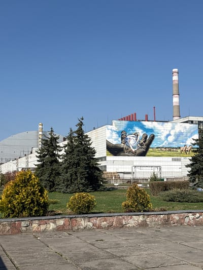 A view from Pryp'yat', Ukraine showing a large building facade decorated with a mural of a hand holding an atomic structure. A smokestack with red and white stripes is visible above the building. The sky is clear blue. A walkway with stone borders and grass is in the foreground. A row of bushes separates the grass from the walkway.