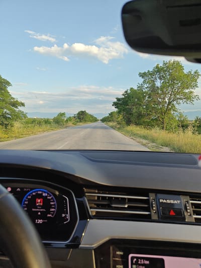 The image captures a view from the interior of a Volkswagen Passat, facing forward through the windshield onto a rural road near Gelemenovo, Bulgaria. The car is traveling at 120 km/h, with an ambient temperature of 25.5°C displayed on the digital instrument cluster, which also shows a song or message with text "Slobodna sam t...". A portion of the infotainment screen is visible, indicating "2.5 km" to a destination. The "PASSAT" branding and a red triangular hazard light button are present on the central dashboard vents.
The asphalt road, marked with a broken white center line and a solid white edge line, extends straight into the distance. Both sides of the road are bordered by overgrown green and dried grass, as well as various deciduous trees and shrubs. In the background, distant mountains or hills are visible beneath a light blue sky with scattered white cumulus clouds. The lighting suggests late afternoon or early evening. The underside of the car's rearview mirror is visible at the top of the frame. No individuals are discernible in the scene.
