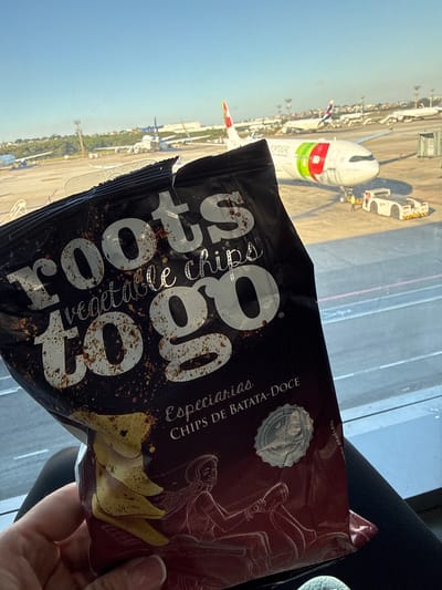 An indoor, eye-level shot captures a person holding an open bag of "roots vegetable chips to go" with an airport tarmac visible through a large window in the background. The foreground is dominated by a person's right hand, revealing only fingers and thumb, holding a dark-colored bag. The bag is open at the top, showing light yellow sweet potato chips within. White text on the bag reads "roots vegetable chips to go," "Especiarias," and "CHIPS DE BATATA-DOCE," with "INDUSTRIA BRASILEIRA" printed vertically along the right edge. A silver circular seal, partially visible, contains text such as "PREMIUM QUALITY" and "REAL FOOD." Below the bag, a segment of the person's dark-clothed leg is visible.

Through the window, the tarmac of Guarulhos Airport in Brazil is brightly lit under a clear blue sky. A white TAP Air Portugal aircraft, featuring green, red, and white livery and the text "PORTUGAL" on its fuselage, is prominently parked. Near this aircraft, two white ground support vehicles with yellow accents are visible, alongside a person wearing a yellow safety vest. In the midground and background, other parked aircraft, airport infrastructure, and a distant line of green trees with utility poles are discernible.