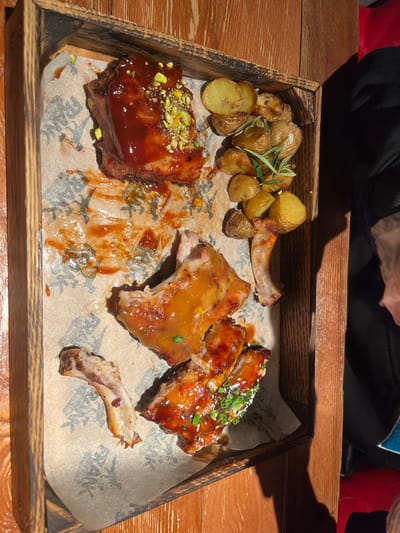 A wooden serving tray, lined with grease-stained parchment paper featuring faint graphic patterns, displays a partially consumed meal. The tray contains several pieces of cooked ribs coated in various sauces; one prominent piece is covered in a dark red sauce topped with green crushed pistachios, while other smaller pieces are sauced and garnished with sesame seeds and chopped green onions. A cleaned rib bone is present, indicative of consumption. Alongside the ribs are multiple small, roasted potatoes, some whole and some halved, garnished with fresh rosemary sprigs. The tray rests on a dark brown wooden surface, possibly a table, with distinct shadows suggesting an overhead light source. To the immediate right of the tray, a dark fabric object and a red object are partially visible. This scene is located in Родниковское сельское поселение, Ukraine.