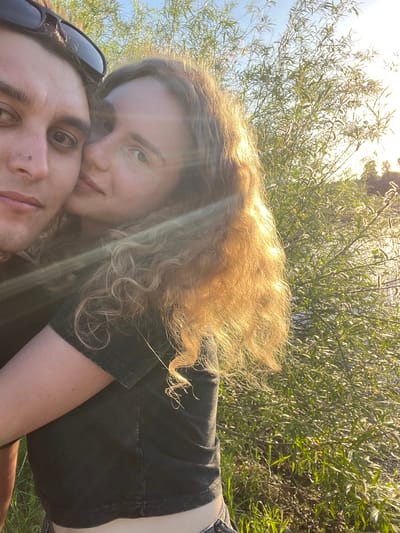 A selfie captures two individuals outdoors, with one person lying on the ground and the other leaning down towards them. The individual on the ground has short, dark hair and is wearing sunglasses perched on their forehead. The person leaning down has long, curly, light brown hair. They are wearing a dark t-shirt. Lush green vegetation forms a backdrop. The exact location within Krasnyi Jar, Russia, is not identifiable from the visual context.