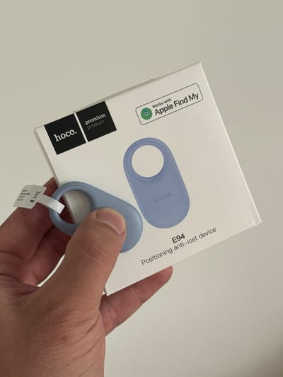 A close-up, high-angle shot in Shirkovo, Russia, presents a human hand holding a white product box and a light blue anti-lost device. The box, branded "hoco." as a "premium product," displays a graphic of the device and features a "Works with Apple Find My" logo. Text on the box identifies the product as "E94 Positioning anti-lost device." The actual light blue device, which is oval-shaped with a circular cutout at one end and "hoco." embossed on its surface, is being held by the thumb and forefinger. A small white pull-tab is partially inserted into the device's circular cutout. The background is a plain, light gray surface.