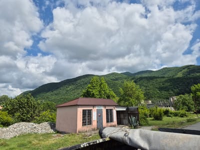 The image shows a small, pink, single-story building with a brown, corrugated metal roof. The building has two windows with metal grates and a central door. Signs are visible on the building. To the left of the building, there is a pile of stones and gravel, and to the right, an agave plant. In the background, lush green mountains rise under a sky with scattered white clouds. Further in the background to the right, an unfinished concrete structure is visible. The foreground shows the edge of a vehicle with a rolled-up tarp. The scene appears to be located in a rural or semi-rural area, consistent with the town of Bzypta, Abkhazia.