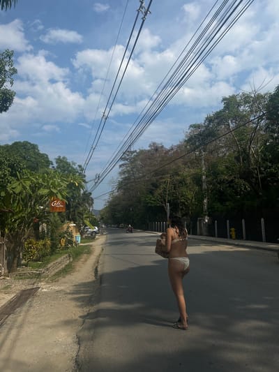 The image shows a woman walking down a paved road in Santa Fe, Costa Rica (latitude: 9.643, longitude: -85.167). The road is lined with lush vegetation on both sides, and the sky is a bright blue with white clouds. There are numerous power lines running above the street. The woman is wearing a white bikini and carrying a brown bag. She is walking away from the camera. Two vehicles are parked on the left side of the road, and two people are riding a scooter on the right side of the road. A brown sign on the left side of the road reads "Cafe."