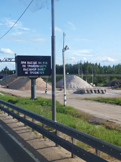 The image shows a digital billboard displaying text in Russian, located in the city of Budovo, Russia. The billboard is mounted on a pole next to a road with a metal guardrail. The text on the billboard reads: "ПРИ ВЪЕЗДЕ НА М11 ПО ТРАНСПОНДЕРУ ВЪЕЗДНОЙ БИЛЕТ НЕ ТРЕБУЕТСЯ" which translates to "UPON ENTERING M11 WITH A TRANSPONDER, AN ENTRY TICKET IS NOT REQUIRED". In the background, there are piles of sand and gravel, a forest, and a bright blue sky with some clouds. The overall scene appears to be a highway toll plaza or an entrance to a toll road. There are no people visible in the image.