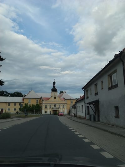 The image shows a street scene in Slavňovice, Czechia. On the left, a large, yellow building with a red roof and a prominent black tower with a clock is visible. The building has multiple windows and appears to be a historical structure. To the right of the street, a row of buildings with white and light gray facades runs along the sidewalk. These buildings have windows and some feature wooden awnings. A red car is parked on the side of the street near the yellow building. The street itself is paved and has white dashed markings. The sky above is partly cloudy, with a mix of blue sky and white and gray clouds. Trees and foliage are visible in the background. The perspective suggests the photo was taken from a vehicle.