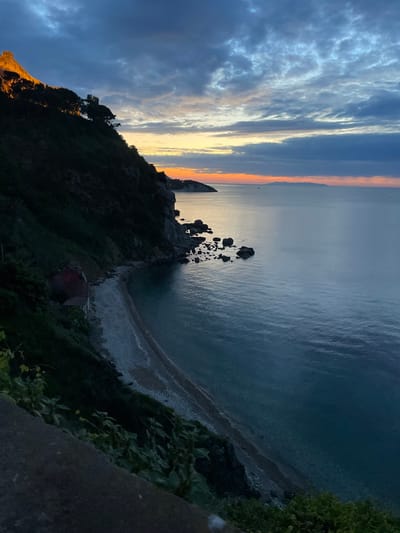 The image shows a view from a cliffside overlooking a small, secluded beach in Portoferraio, Italy. The coordinates (latitude: 42.817, longitude: 10.332) pinpoint the location to the western part of the island of Elba, specifically near the Forte Stella, a historical fort. 

The scene captures the calmness of twilight, with a clear, blue sky fading into a soft orange horizon. The sea is a deep blue, reflecting the colors of the sky, and stretches out towards the distant horizon. The beach below is pebbled and curves gently along the coastline. The cliffside is covered in dense greenery and shadowed by the approaching darkness. Several large rocks are visible in the shallow water near the shore.  The image is taken from a high vantage point on the cliff, providing a wide, panoramic view of the landscape.  There are no people or objects in the foreground, suggesting that the scene is empty and peaceful.