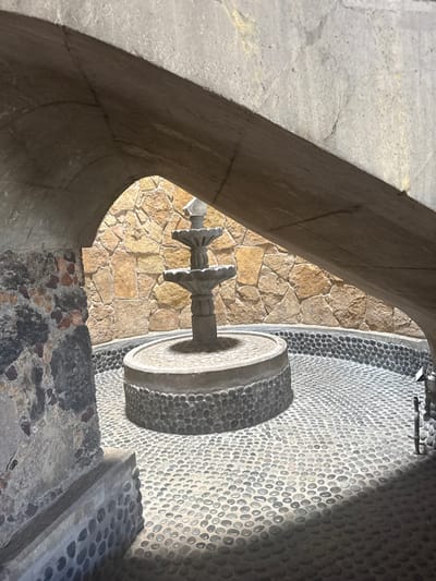 The image shows an outdoor fountain viewed through an arched opening, possibly a window or doorway. The fountain is made of stone and features a circular basin. The walls surrounding the fountain are constructed of stone, with the immediate background consisting of light-colored stone blocks, and a wall or surface paved with small, round pebbles. The arch framing the view is also made of stone. The location is Tres de Mayo, Mexico.