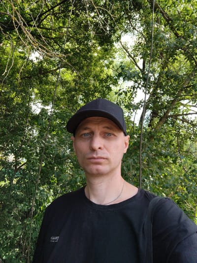 A man wearing a black baseball cap and a black t-shirt is standing outdoors. He has light skin and blue eyes, and is looking directly at the camera. The man is wearing a silver chain necklace. Behind him, there is dense green foliage from trees and some kind of fencing. The overall environment appears to be a natural setting, possibly a park or a backyard, in Mokshan, Russia. The lighting suggests it is daytime. There are no other people or animals visible in the frame.