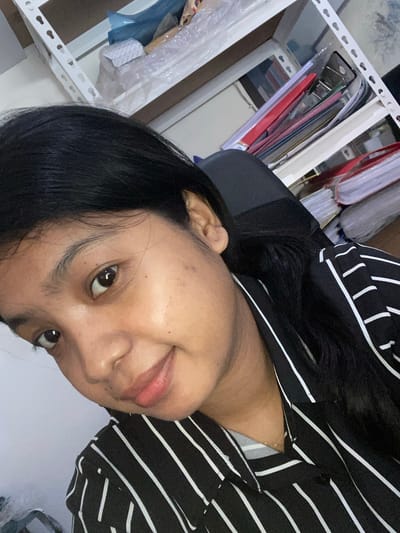 A close-up, rotated selfie shows a woman indoors. She has dark hair and appears to be wearing a black and white striped shirt. In the blurred background, a white shelving unit holds files and documents. This image provides no information about Negara, Indonesia.