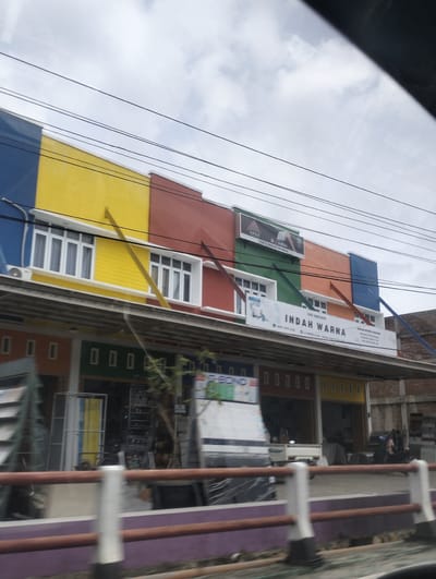 The image shows a multi-story building with a brightly colored facade in the city of Ujong Duabelas, Indonesia. The building features a red, orange, green, yellow, and blue color scheme. The upper level has several windows with white frames. A prominent sign on the building reads "INDAH WARNA" and appears to be for a paint or color store, with the tagline "TOKO INDAH WARNA". Below the upper level, the ground floor consists of multiple shopfronts. In front of the building, a street or sidewalk is visible with a railing. The sky is overcast. Various vehicles, including a white truck, a motorcycle, and possibly a car, are parked or in motion near the building. There are a few people visible in the distance, some on foot and some on a motorcycle. The scene suggests a commercial area on a cloudy day.