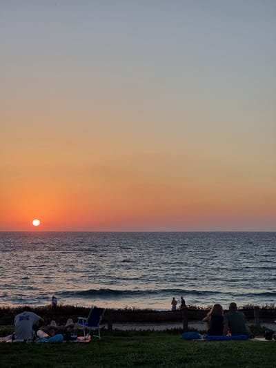 The image depicts a sunset at Palmachim Beach, Israel. A couple is seated on the grass, facing the ocean and the setting sun. The sun, a bright orb, is positioned near the horizon, casting a warm orange glow across the sky and the water. Gentle waves roll towards the shore, where a few other figures can be seen near the water's edge. In the foreground, several blankets and a blue beach chair are laid out on the grass. The scene is serene and picturesque, capturing the beauty of a coastal sunset.