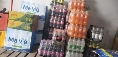 The image showcases a stockpile of beverages. There are stacks of bottled sodas in various flavors, packaged in plastic wrap, including Coca-Cola, Fanta, and Sprite. Cardboard boxes labeled "Ma Vie" are also present. Above them are boxes of "Sana" fruit juice. A collection of crates is visible to the left.