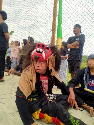 This image captures an outdoor scene in Tamansari, Indonesia, featuring individuals engaged in what appears to be a cultural performance or event, separated from spectators by a chain-link fence.
In the central foreground, a person is seated on a light-colored paved surface, facing slightly right and forward. This individual wears an elaborate red mask with fangs and dark, shaggy hair. Their visible face beneath the mask features eyes painted white with red streaks descending from them, and their mouth is slightly open. They are dressed in a black long-sleeved shirt with a red graphic, a light brown tattered shawl draped over their shoulders, black pants, and red and yellow fringed adornments around their ankles. Their left arm is extended forward, resting on the ground.
To the immediate right of this figure, an older man is also seated on the ground, looking forward. He wears a dark cap, a dark t-shirt with the partial text "THNT POR" visible, and dark pants. He holds a light-colored cylindrical object in his right hand.
A chain-link fence runs across the mid-ground, separating the foreground subjects from a group of spectators. Behind the fence, a crowd of people, including several women, some wearing hijabs, are observing. One woman in a light pink top smiles and gestures with her left hand. Further right, a man with a dark cap and long-sleeved shirt stands with his arms crossed, looking to the right. Two vertical posts, one wrapped in green and yellow fabric, are visible directly behind the fence. The background consists of an overcast sky and distant green vegetation. Small green plants grow on the paved surface in the foreground.