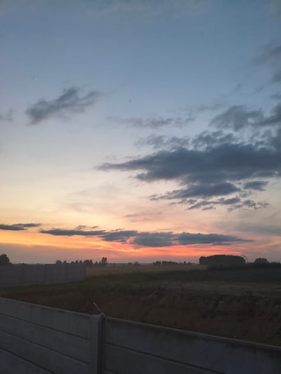 This image depicts a rural landscape at dusk, likely in or near Galonki, Poland. The sky transitions from a pale blue at the top to vibrant hues of orange and pink near the horizon, indicating a sunset. Dark, scattered clouds drift across the sky. In the foreground, a concrete fence, constructed from modular panels, runs horizontally across the frame, suggesting an area under construction or a boundary. Beyond the fence, a field is visible, with darker areas of soil and lighter patches that could be vegetation or tilled land. A line of trees forms a distant treeline, and a few structures, possibly buildings or agricultural elements, are faintly discernible in the far distance. The overall scene is serene and devoid of human presence.