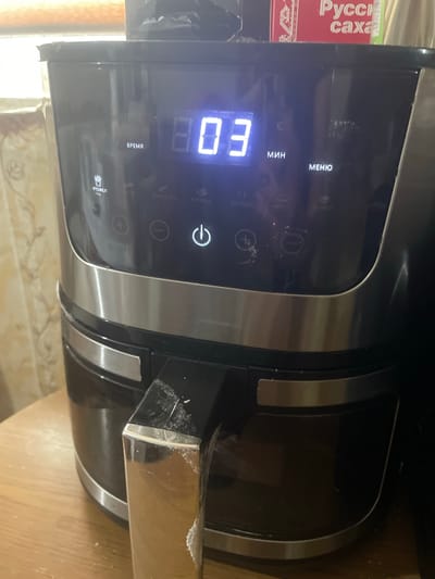 This media file shows a close-up of a black air fryer with a digital display. The display shows "03" in large, illuminated blue digits, likely indicating minutes remaining in a cooking cycle. Above the digits, the word "ВРЕМЯ" (TIME) is visible in Russian. To the left of the display, a red and white box with Russian text, "Русская сахара" (Russian sugar), is partially visible.
The air fryer has several buttons and icons. There's a power button with a circular arrow symbol, and buttons with "+" and "-" symbols for adjusting settings. The words "МЕНЮ" (MENU) and "МИН" (MIN) are also visible in Russian, suggesting menu selection and minute adjustments. Below the "ВРЕМЯ" display, there's an icon of a plate with food and the word "РТОФЕЛ" (POTATO), indicating a preset cooking mode. Other faint text, possibly other preset modes like "КУРИЦА" (CHICKEN), is also visible.
The air fryer appears to be on a wooden surface, likely a kitchen countertop. The lighting suggests it is indoors, with no specific indication of the time of day or weather. The scene is a still shot, with no activity or event taking place other than the air fryer's display indicating a countdown. There are some smudges and minor damage visible on the air fryer's surface, particularly around the display and on a metal handle-like component on the right side. The overall impression is a functional kitchen appliance in use or ready for use.