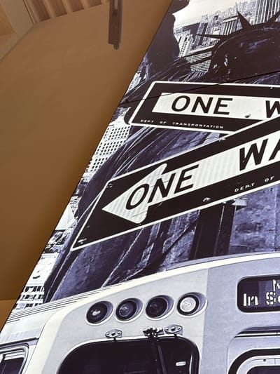 This media file is a photograph of a large, angled display or mural. The display features a black and white image with a gritty, urban aesthetic. Prominently displayed are two "ONE WAY" street signs, overlapping each other and angled towards the viewer. The signs are bold and stark, with white lettering on a black background, and "DEPT. OF TRANSPORTATION" is printed vertically along the side of one sign.

Behind the signs, a silhouette of the Statue of Liberty is visible, rendered in dark tones against a backdrop of what appears to be a city skyline, also in black and white. To the right of the signs is a close-up view of what seems to be part of a vintage vehicle or train interior, with a series of circular buttons or dials and a digital display. The overall impression is one of a dynamic, possibly historical, New York City scene.

The lighting in the image is bright, suggesting daytime, but the grayscale treatment of the mural creates a timeless, almost noir-like atmosphere. The image is presented at an angle, indicating it's a photograph of a physical display that is not perfectly perpendicular to the camera. There are no people visible in the image. The context provided, Mülheim-Kärlich, Germany, is external information and not directly discernible from the visual content of the photograph itself, which strongly suggests a US urban environment.