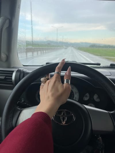 A person's hand with multiple rings is resting on the steering wheel of a Toyota vehicle. The vehicle appears to be driving on a wet road, as suggested by the droplets on the windshield and the visibility conditions. The surrounding environment is blurred, indicating motion. It is not possible to confirm the location in the city of Чистенское сельское поселение, Ukraine, from this image.