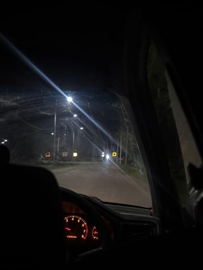 The image is taken from the interior of a car at night, looking out through the windshield. The scene depicts a dimly lit road with streetlights and the headlights of other vehicles. In the foreground, the silhouette of a person's head is visible on the left, and the dashboard with a lit tachometer is visible on the lower left. The road appears to be paved and wet, with reflections of lights on its surface. Several utility poles with lights are spaced along the side of the road. Further down the road, there are illuminated signs and buildings. The surrounding environment appears to be a rural or suburban area with trees visible on the right side of the frame. The image is grainy and low-light, characteristic of night photography. The location is specified as Ptichnik, Russia.