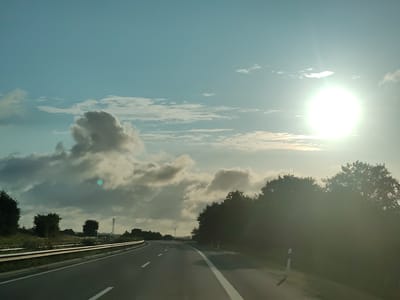 The image captures a view from a vehicle traveling on a highway in Gremersdorf, Germany. The sun is prominently displayed in the upper right corner of the frame, partially obscured by clouds, casting a bright glare across the scene. The sky is a clear blue with scattered white and grey clouds, some of which are large and puffy. On the left side of the highway, a metal guardrail runs alongside a grassy verge with trees. The road itself is asphalt, marked with white lane lines. On the right side, a dense line of trees forms a dark silhouette against the bright sky. In the distance, a wind turbine can be seen against the cloudy horizon. The overall impression is of a scenic highway drive under a partly cloudy sky.
