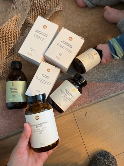 The scene is described as an indoor setting in Waldgrehweiler, Germany, though this specific geographical location is not visually verifiable from the image content. The image depicts various nutritional supplement products from the "Sunday Natural" brand, interacting with human hands, arranged on a floor composed of a patterned carpet and adjacent wooden planks.
**Environment:** The floor surface in the foreground and center consists of a light-colored, patterned carpet, predominantly beige with subtle horizontal stripes in shades of light pink and brown. To the lower right and extending upwards, a light brown wooden floor is visible. Crumpled brown honeycomb-patterned paper packaging material is present in the upper-left corner of the carpet, indicating recently opened packages.
**Objects:** Seven "Sunday Natural" brand products are visible. These include three white cardboard boxes and four amber glass bottles with black screw caps, all featuring light-colored labels with the brand's lotus logo:
1. **SUNDAY ESSENTIALS MULTI-AMINOS PLUS:** A white cardboard box (120 Kapseln, vegan).
2. **NOBLE STRONG MUSHROOM COFFEE:** A white cardboard box (80g, vegan).
3. **ALL IN ONE ONE A DAY:** A white cardboard box (60 Kapseln).
4. **ALL IN ONE LIPOSOMAL:** An amber glass bottle (250ml, vegan).
5. **GLYCIN 500:** An amber glass bottle (500mg, 500 Kapseln, vegan), positioned horizontally.
6. **OMEGA 3 COMPLETE DHA: EPA: ALA:** An amber glass bottle (90 Softgel Kapseln, vegan), positioned horizontally.
7. **MAGNESIUMGLYCINAT DEPOT:** An amber glass bottle (120 Kapseln á 765 mg, 150 mg Magnesium, vegan).
**People, Actions, and Interactions:**
1. An adult's left hand, exhibiting a light skin tone, holds the "MAGNESIUMGLYCINAT DEPOT" bottle in the lower-center foreground.
2. A young child's right hand, partially obscured and wearing a blue, green, and white striped sleeve, grasps the "GLYCIN 500" bottle in the upper-right.
3. A young child's bare left foot and lower leg, clad in light grey pants, are visible in the upper-right quadrant of the carpet.
The scene indicates an interaction where both an adult and a child are in contact with the recently unboxed supplement products.
