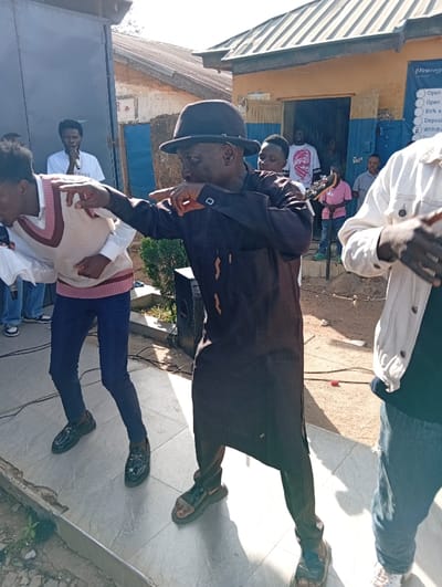 The image depicts an outdoor scene in Jebbu Miango, Nigeria, showing a group of individuals gathered, with several engaged in performance. In the foreground, a man wearing a dark fedora, a dark brown long-sleeved tunic (agbada-style), and dark sandals stands with his right arm extended forward and fingers clasped, indicating a pose or dance move. To his left, another young man, dressed in a white collared shirt, a beige and pinkish V-neck sweater vest, dark trousers, and black loafers, is bent forward with both arms raised, also appearing to dance. Partially visible on the far right, a third individual wears a light-colored jacket and blue jeans, with their hands positioned in a dynamic gesture.
In the background, positioned behind the left dancer, a man in a white T-shirt holds a microphone. Multiple other individuals are visible observing from a doorway and around a building with a corrugated metal roof. This building features light brown walls with blue trim around an open doorway, and a partially visible sign on the wall, reading "Firstmo..." with bullet points underneath, likely indicating banking services. To the left of the main performers, a large black speaker box is present. The foreground ground surface consists of light-colored tiles, beyond which is a dirt or gravel area with some visible cables. The lighting indicates a bright, sunny daytime environment.