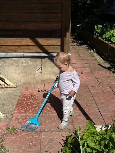 A young child with blonde hair, wearing a light purple long-sleeved shirt with a floral pattern and white pants with red hearts, stands on a red tiled pathway next to a weathered wooden building in Дзержинец-83 Снт, Russia. The child is holding a bright blue broom with gray bristles, appearing to be sweeping. Sunlight casts a shadow from the building onto the pathway and the child. To the right of the pathway, there is a low, dark planter box filled with lush green plants and foliage. The ground is cracked in some areas, and there is debris on the pathway.
