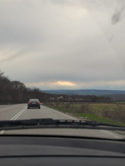 The image shows a view from inside a moving vehicle, likely a car, looking forward onto a road in Buinovo, Bulgaria. The sky is overcast, with a bright patch of light breaking through the clouds. In the distance, a hazy landscape with fields and hills can be seen. The road has two lanes marked by a double white line. A black van is directly ahead, traveling in the same direction. The foreground includes the vehicle's windshield wiper and the interior side of the car.